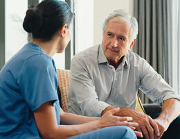  A man in street clothes sits in a hallway, talking to a woman in scrubs.